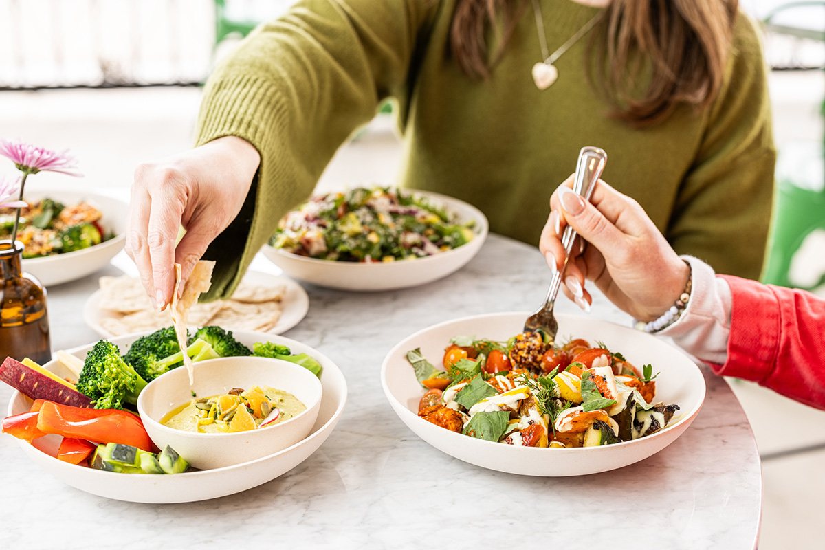 Three healthy dishes on a round whit table at Flower Child restaurant with people reaching in with fork ready to eat.