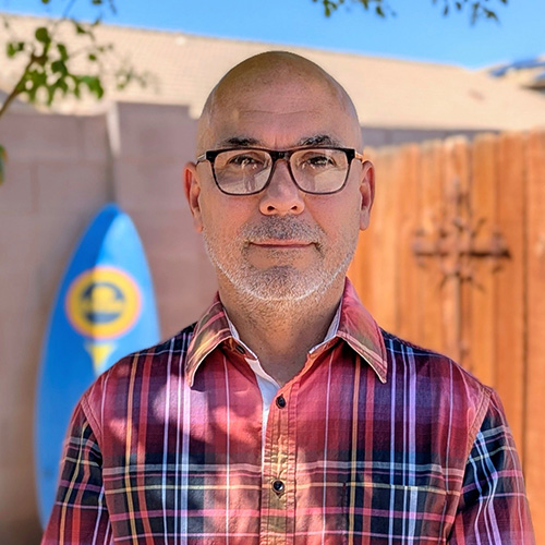 Man wearing a red and blue plaid collared shirt standing in from of a brick wall with a light blue surf board leaning against the wall.